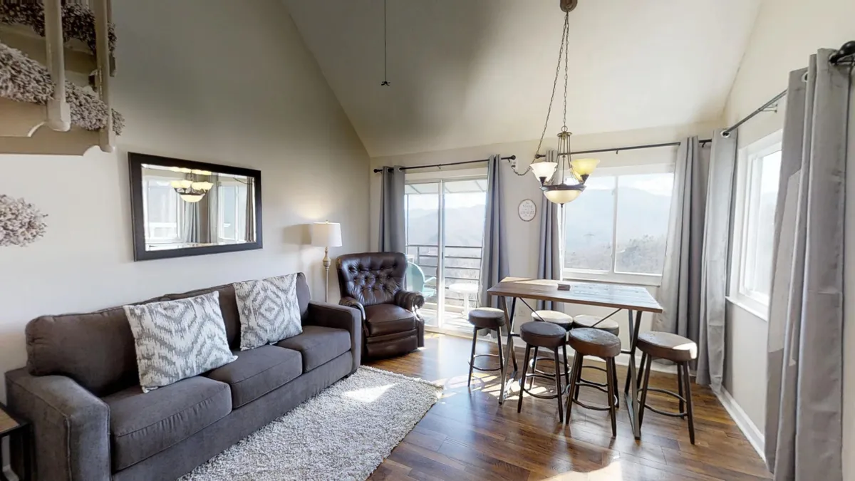 A view of the living area with a plush sofa adorned with decorative pillows, a dining table with stools, and large windows that offer a glimpse of the scenic outdoors. The natural light floods the space, creating a warm, welcoming atmosphere that aligns with the narrative's emphasis on comfort and view.