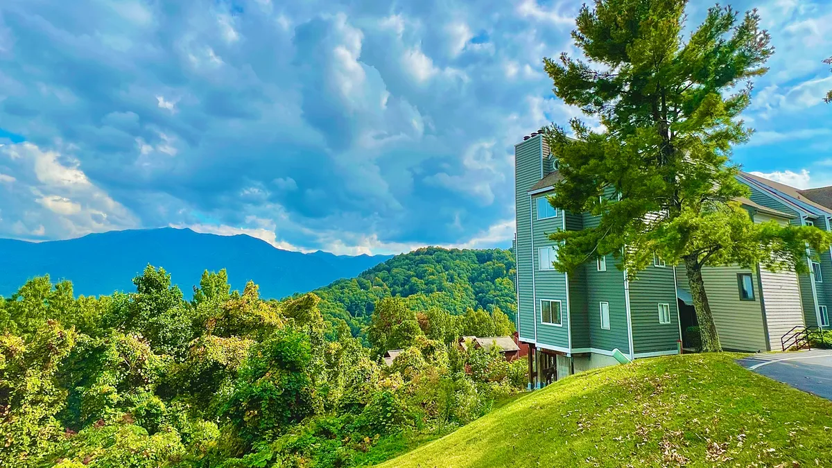 The hero image showcases a modern condo exterior nestled amid lush greenery with panoramic, unobstructed views of the Great Smoky Mountains and Mt. LeConte. The scene is peaceful, with golden sunset light spilling over the mountain ridges, inviting you to relax outdoors and take in the natural beauty.