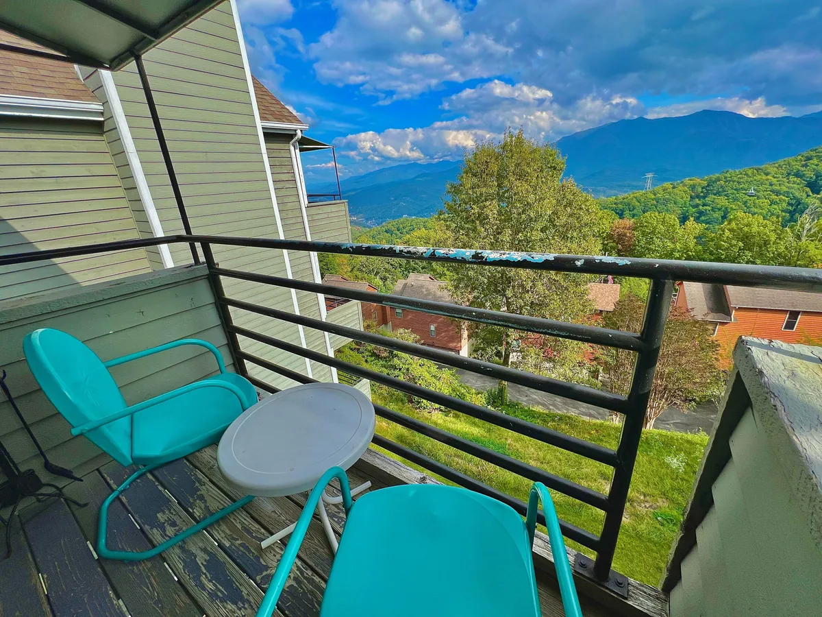 An exterior shot focusing on the balcony, featuring turquoise chairs and a small table set against a stunning mountain vista. The balcony invites guests to relax, enjoy a coffee, or socialize while soaking in the views of the Smokies and Mt. LeConte.
