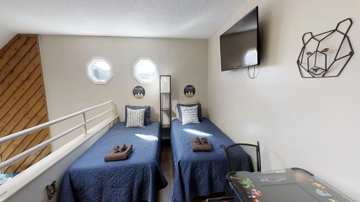 A playful loft area furnished with two twin beds dressed in blue bedspreads, bear-themed wall decor, and a TV mounted on the wall. The space is clearly designed with families or children in mind, offering both relaxation and play.