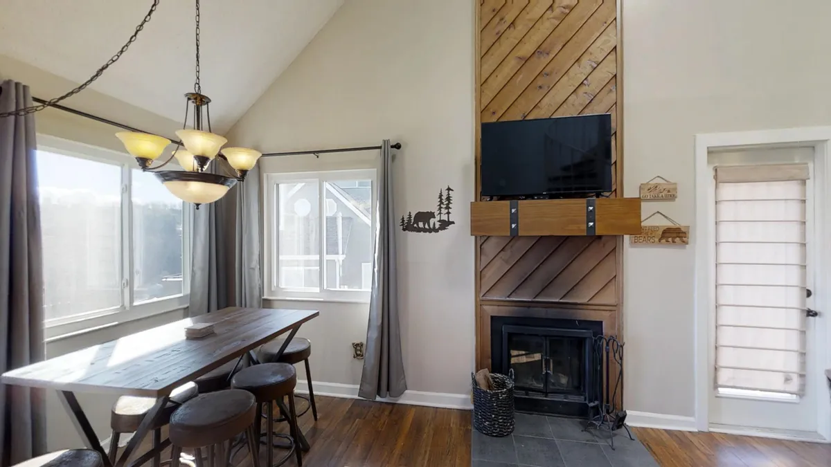 This image presents the living room area, showcasing a dining table, a fireplace, a flat-screen television, and large windows that allow natural light to brighten the space. The seating arrangement is inviting, hinting at comfort and togetherness, and the fireplace is an eye-catching focal point.
