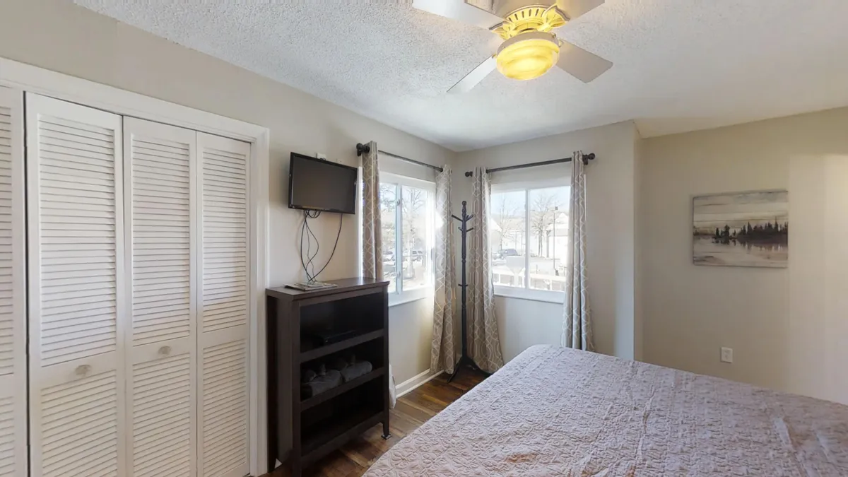 A cozy bedroom with a comfortable bed, ample windows letting in sunlight, a functional dresser, a television, and accent artwork. The space feels homey and relaxed, ideal for guests seeking comfort and leisure.