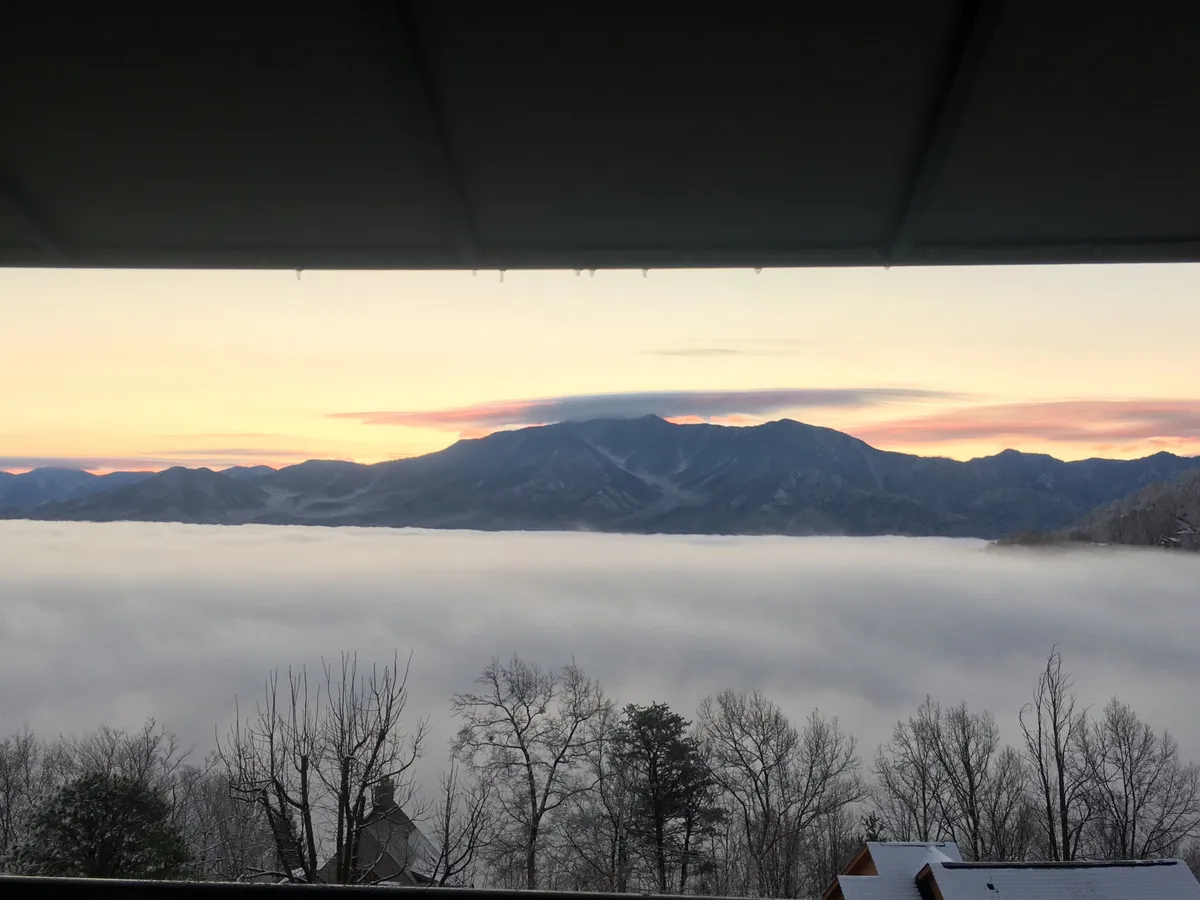A stunning view of a mountain range with fog rolling through the valley and a colorful sunset casting warm hues across the sky. The tranquil scenery emphasizes the beauty and serenity of the location, inviting guests to unwind and take in the majestic surroundings.