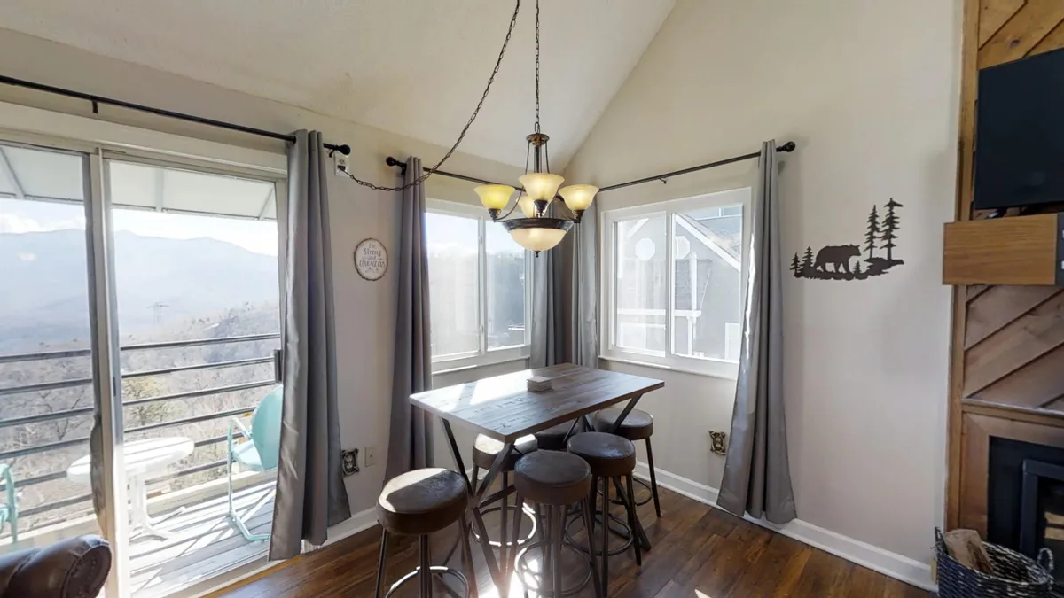 A relaxed and inviting dining area featuring a sturdy table set for six, illuminated by a chandelier. Large windows frame sweeping mountain views, and a bear silhouette adorns the wall, creating a cozy mountain ambiance. The natural light and scenic backdrop make this space perfect for family meals and gatherings.