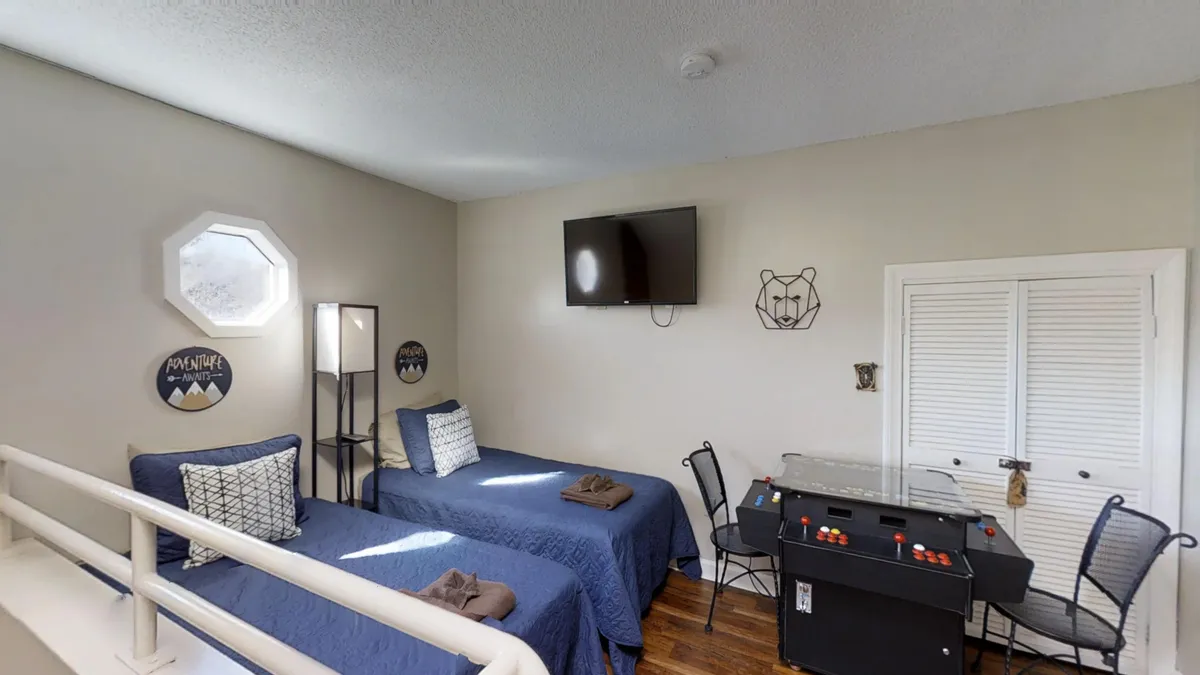 An interior scene featuring two twin beds with blue bedspreads, a small dining table and chairs, a wall-mounted TV, and playful decor. The setup is ideal for group activities like playing games, watching movies, or casual group meals.