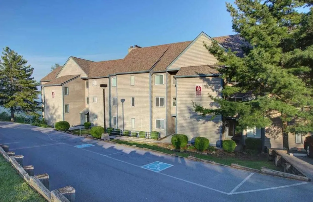 This image captures the exterior of the building, featuring the welcoming facade, landscaped shrubs, and a parking area. The overall mood is inviting, with visible balconies that hint at spaces perfect for gathering and relaxing with friends.